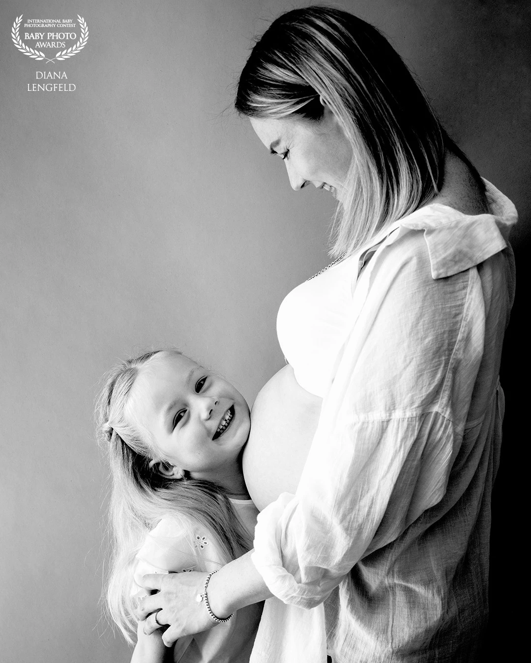 Schwarz-Weiß: Ein Mädchen schmiegt sich lachend an den Babybauch ihrer Mutter; mit Baby Photo Awards Signet