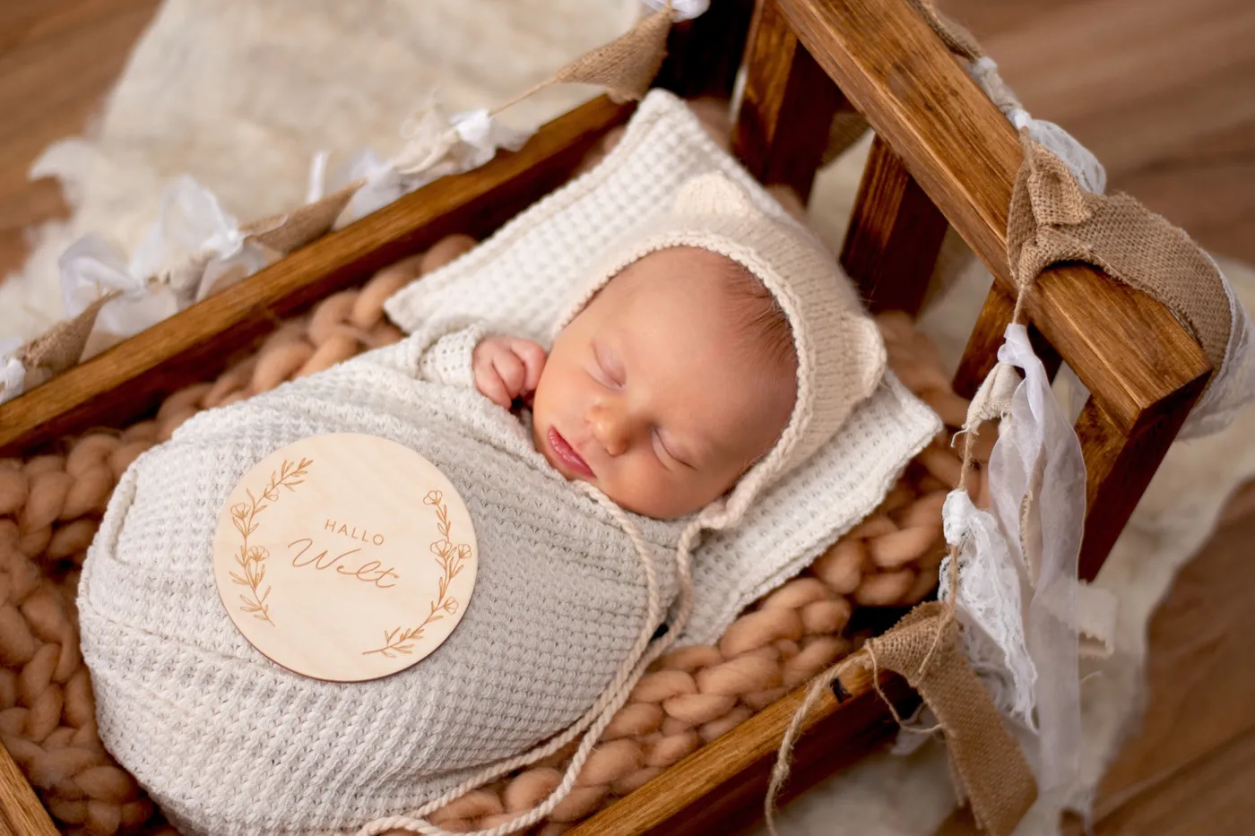 Schlafendes Baby mit Bärchenmütze in einer Holzwiege, auf der Decke liegt eine Holzscheibe mit der Aufschrift „Hallo Welt“